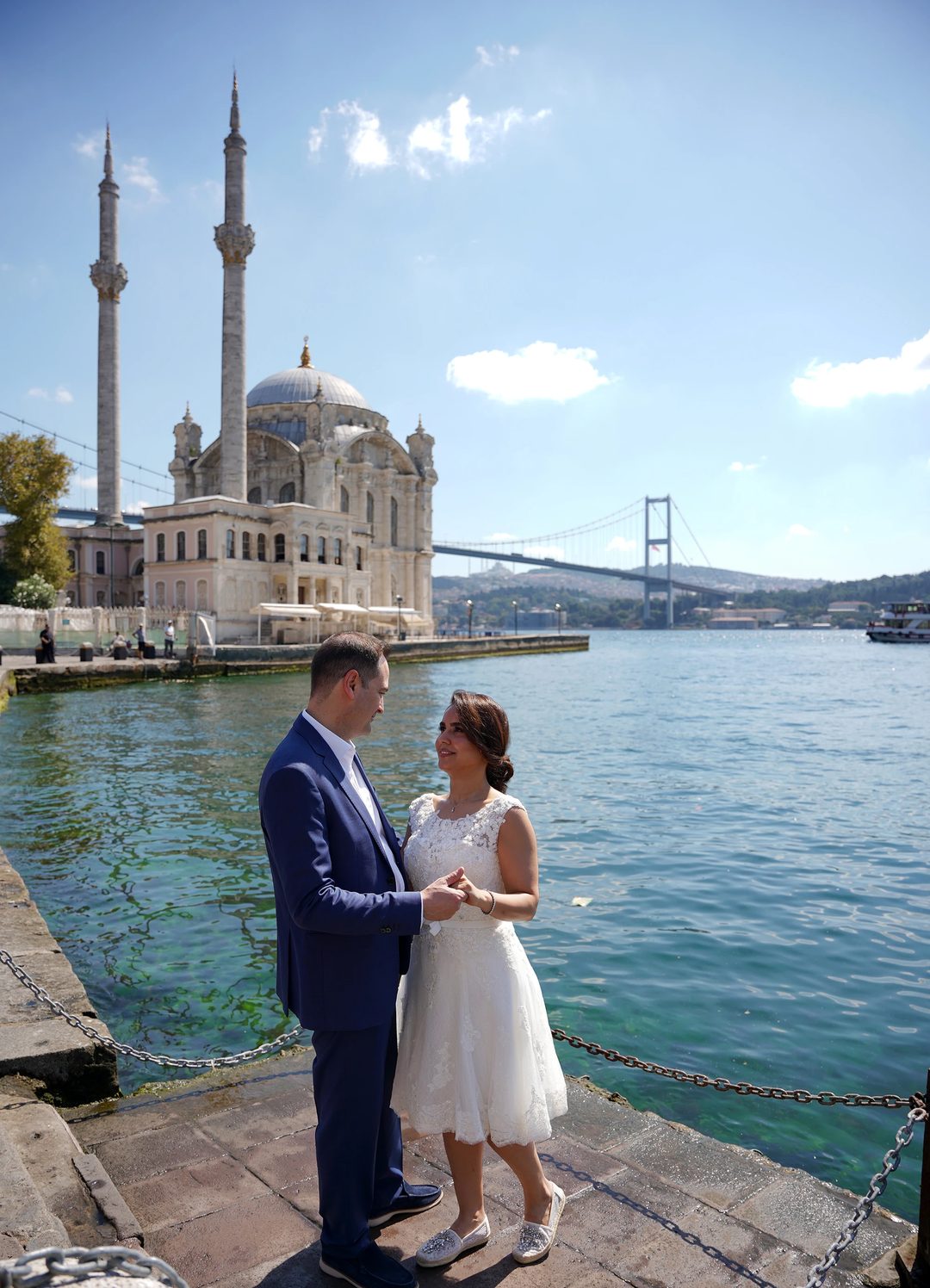 Setayesh and Aydin holding hands on the waterfront, with the Ortakoy Mosque and the Bosphorus Bridge behind them.